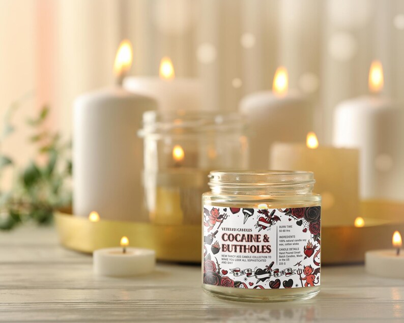 Może przedstawiać: Świeca w szklanym słoiku z etykietą z napisem "Cocaine & Buttholes" z dekoracyjnym wzorem. Świeca otoczona jest zapalonymi świecami o r&oacute;żnych rozmiarach i złotą tacą. Tło jest rozmyte.
