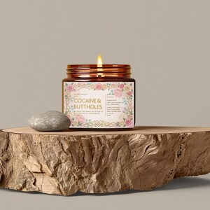 Pu&ograve; includere: Una candela accesa in un barattolo di vetro con un'etichetta floreale che recita "Cocaine & Buttholes". Il barattolo &egrave; posto su una lastra di legno accanto a una piccola pietra grigia. La candela ha un coperchio marrone.