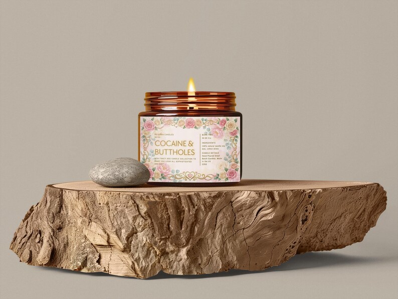 Pu&ograve; includere: Una candela accesa in un barattolo di vetro con un'etichetta floreale che recita "Cocaine & Buttholes". Il barattolo &egrave; posto su una lastra di legno accanto a una piccola pietra grigia. La candela ha un coperchio marrone.