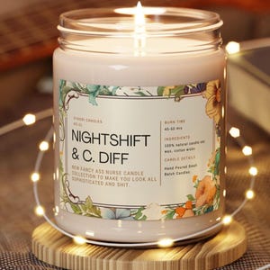 May include: A lit candle in a glass jar with a label that reads "NIGHTSHIFT & C. DIFF." The label has floral accents and text indicating it's a soy wax candle with a 45-50 hour burn time. The candle sits on a wooden coaster, surrounded by fairy lights.