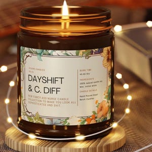 May include: A lit brown glass jar candle with a label that reads "DAYSHIFT & C. DIFF." The label includes floral designs and text about the candle's burn time and ingredients. The candle sits on a wooden coaster, surrounded by string lights.