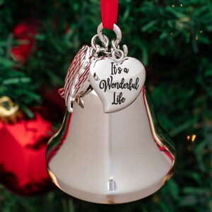 May include: Silver bell Christmas ornament with a red ribbon. The ornament has a heart-shaped charm that reads "It's a Wonderful Life." A decorative wing is attached to the top of the bell. The ornament is hanging on a Christmas tree.