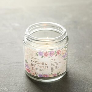 May include: A clear glass jar candle with a lit wick. The label reads "COCAINE & BUTTHOLES" surrounded by a floral design in pink, purple, and cream colors. The candle is on a gray surface.