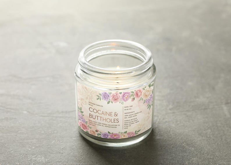 May include: A clear glass jar candle with a lit wick. The label reads "COCAINE & BUTTHOLES" surrounded by a floral design in pink, purple, and cream colors. The candle is on a gray surface.