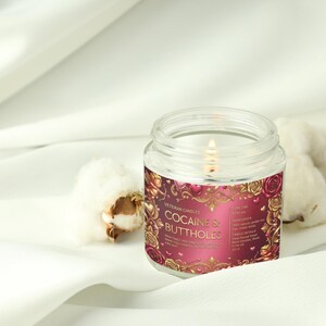 Peut inclure: Une bougie en pot en verre transparent allum&eacute;e avec une &eacute;tiquette qui dit "COCAINE & BUTTHOLES" en lettres dor&eacute;es. L'&eacute;tiquette a un fond bordeaux avec des accents floraux dor&eacute;s. Deux boules de coton sont &agrave; c&ocirc;t&eacute; de la bougie sur une surface blanche.