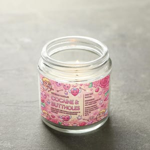 May include: A clear glass jar candle with a pink label featuring cartoon cupids, hearts, and roses. The label reads "COCAINE & BUTTHOLES" in bold letters. The candle is lit, with a small flame visible.