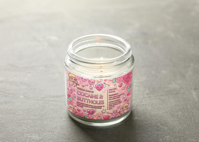 May include: A clear glass jar candle with a pink label featuring cartoon cupids, hearts, and roses. The label reads "COCAINE & BUTTHOLES" in bold letters. The candle is lit, with a small flame visible.