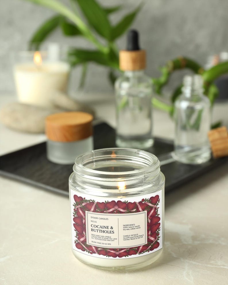 May include: A lit, clear glass jar candle with a label that reads "Cocaine & Buttholes" surrounded by a heart design. The candle is on a white surface with other blurred apothecary bottles and a candle in the background.