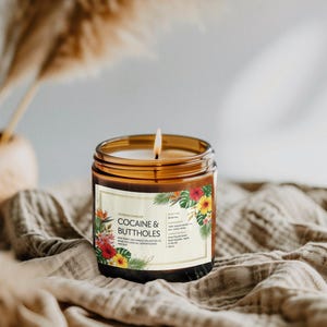 May include: A lit candle in a brown glass jar with a label that reads "COCAINE & BUTTHOLES". The label features tropical flowers and leaves. The candle sits on a tan cloth.