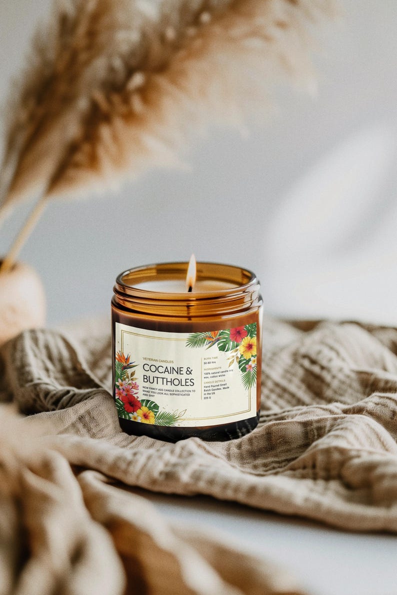 May include: A lit candle in a brown glass jar with a label that reads "COCAINE & BUTTHOLES". The label features tropical flowers and leaves. The candle sits on a tan cloth.