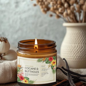 May include: A lit brown glass jar candle with a label featuring floral designs and the text "COCAINE & BUTTHOLES". The candle is surrounded by vanilla beans, cinnamon sticks, and a vase of dried flowers.