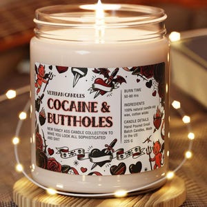 Może przedstawiać: Zapalone, kremowe świeca w szklanym słoiku z etykietą z napisem "COCAINE & BUTTHOLES". Etykieta zawiera ilustracje w stylu tatuażu przedstawiające serca, r&oacute;że i diabły. Świeca stoi na drewnianej podstawce, otoczona łańcuchem świetlnym.