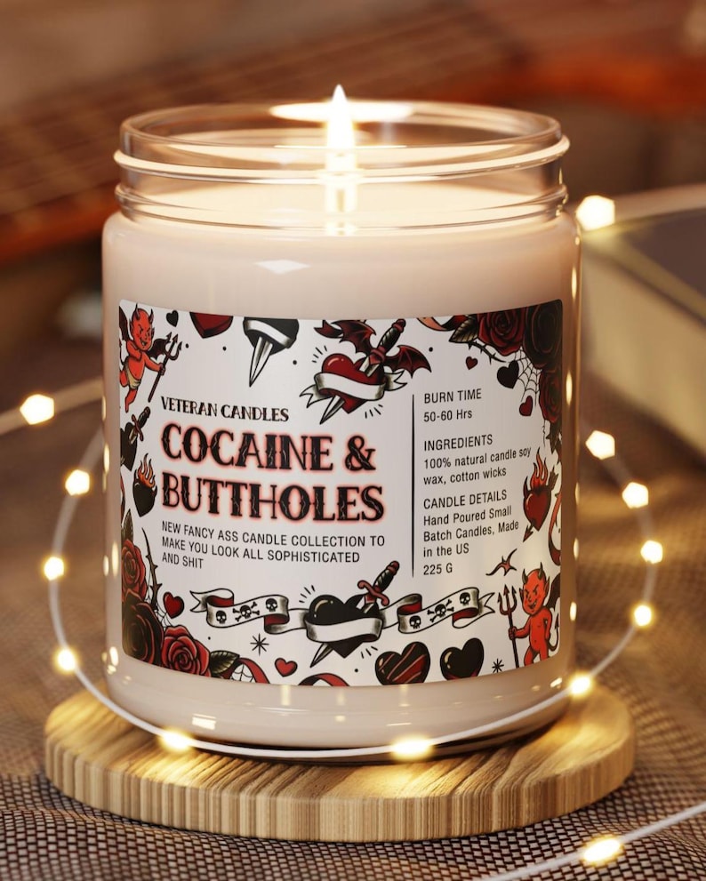 Może przedstawiać: Zapalone, kremowe świeca w szklanym słoiku z etykietą z napisem "COCAINE & BUTTHOLES". Etykieta zawiera ilustracje w stylu tatuażu przedstawiające serca, r&oacute;że i diabły. Świeca stoi na drewnianej podstawce, otoczona łańcuchem świetlnym.