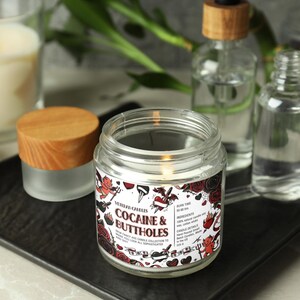 Może przedstawiać: Zapalone świeca w szklanym słoiku z drewnianą pokrywką, z napisem "Cocaine & Buttholes" z czerwonym i czarnym wzorem. Inne szklane butelki i zapalona świeca są w tle. Świeca stoi na czarnej tacy.