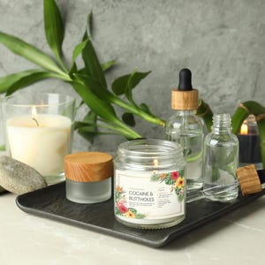 May include: A lit candle in a glass jar with a wooden lid, and a label that reads "COCAINE & BUTTHOLES" with floral designs. The candle sits on a black tray with other glass bottles and a lit candle in a glass.