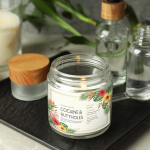 May include: A lit glass jar candle with a wooden lid, labelled "COCAINE & BUTTHOLES" with floral graphics. Other glass bottles and a lit candle are in the background. The candle sits on a black tray.