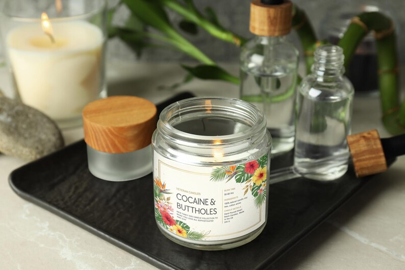 May include: A lit glass jar candle with a wooden lid, labelled "COCAINE & BUTTHOLES" with floral graphics. Other glass bottles and a lit candle are in the background. The candle sits on a black tray.