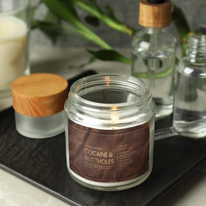 May include: A lit candle in a clear glass jar with a brown label that reads "COCAINE & BUTTHOLES". Other items include a small jar with a wooden lid, two glass bottles with wooden stoppers, and a lit candle in a glass.