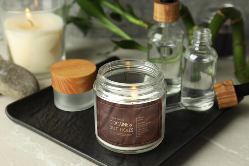 May include: A lit candle in a clear glass jar with a brown label that reads "COCAINE & BUTTHOLES". Other items include a small jar with a wooden lid, two glass bottles with wooden stoppers, and a lit candle in a glass.