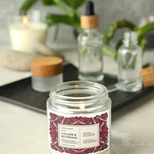 Puede incluir: Una vela encendida en un frasco de vidrio transparente con una etiqueta que dice "Cocaine & Buttholes" rodeada por un dise&ntilde;o de coraz&oacute;n. La vela est&aacute; sobre una superficie blanca con otras botellas de boticario borrosas y una vela en el fondo.