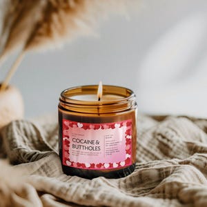 May include: A lit candle in a clear glass jar with a brown label that reads "COCAINE & BUTTHOLES" in pink and white text. The label is decorated with red and pink hearts. The candle sits on a tan cloth.