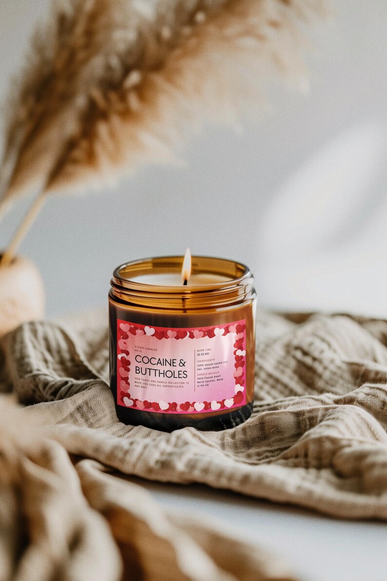 May include: A lit candle in a clear glass jar with a brown label that reads "COCAINE & BUTTHOLES" in pink and white text. The label is decorated with red and pink hearts. The candle sits on a tan cloth.