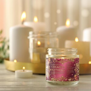 Peut inclure: Une bougie en pot allum&eacute;e avec une &eacute;tiquette qui dit "Cocaine & Buttholes" est au centre, entour&eacute;e d'autres bougies allum&eacute;es de tailles vari&eacute;es. L'&eacute;tiquette est rose et dor&eacute;e avec des motifs floraux. L'arri&egrave;re-plan est doucement &eacute;clair&eacute;.