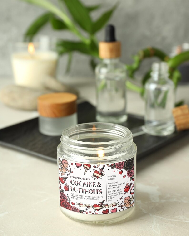 May include: A lit, clear glass jar candle with a label that reads "COCAINE & BUTTHOLES" with a design of roses, hearts, and cherubs. The candle is part of the "VETERAN CANDLES" collection. Other glass jars and a black tray are in the background.