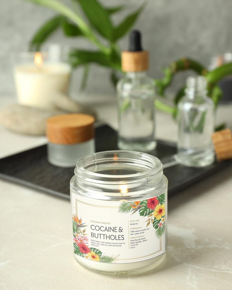 May include: A lit, clear glass jar candle with a label that reads "COCAINE & BUTTHOLES" with a floral design. Other glass bottles and a candle are in the background. The candle is part of the "NEW FANCY AND CANDLE COLLECTION TO MAKE YOU LOOK ALL SOPHISTICATED AND SASSY."