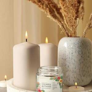 May include: A still life composition with lit candles, a glass jar candle with the text "COCAINE & BUTTHOLES", and a vase with dried plants. The candles are white and cylindrical, set on a terrazzo tray. The jar candle has a floral design.