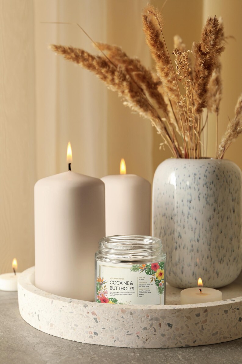 May include: A still life composition with lit candles, a glass jar candle with the text "COCAINE & BUTTHOLES", and a vase with dried plants. The candles are white and cylindrical, set on a terrazzo tray. The jar candle has a floral design.