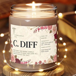 May include: A lit candle in a glass jar with a label that reads "C. DIFF." The label includes floral designs and text: "NEW FANCY ASS NURSE CANDLE COLLECTION TO MAKE YOU LOOK ALL SOPHISTICATED AND SHIT." The candle sits on a wooden coaster.