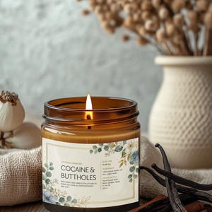 May include: A lit brown glass jar candle with a label that reads "COCAINE & BUTTHOLES". The candle is part of the Veteran Candles collection. The candle is surrounded by cinnamon sticks, vanilla beans, and a vase with dried flowers.