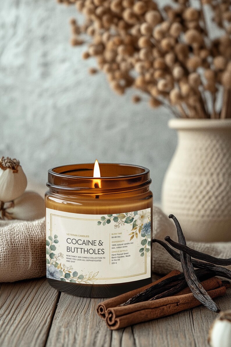 May include: A lit brown glass jar candle with a label that reads "COCAINE & BUTTHOLES". The candle is part of the Veteran Candles collection. The candle is surrounded by cinnamon sticks, vanilla beans, and a vase with dried flowers.