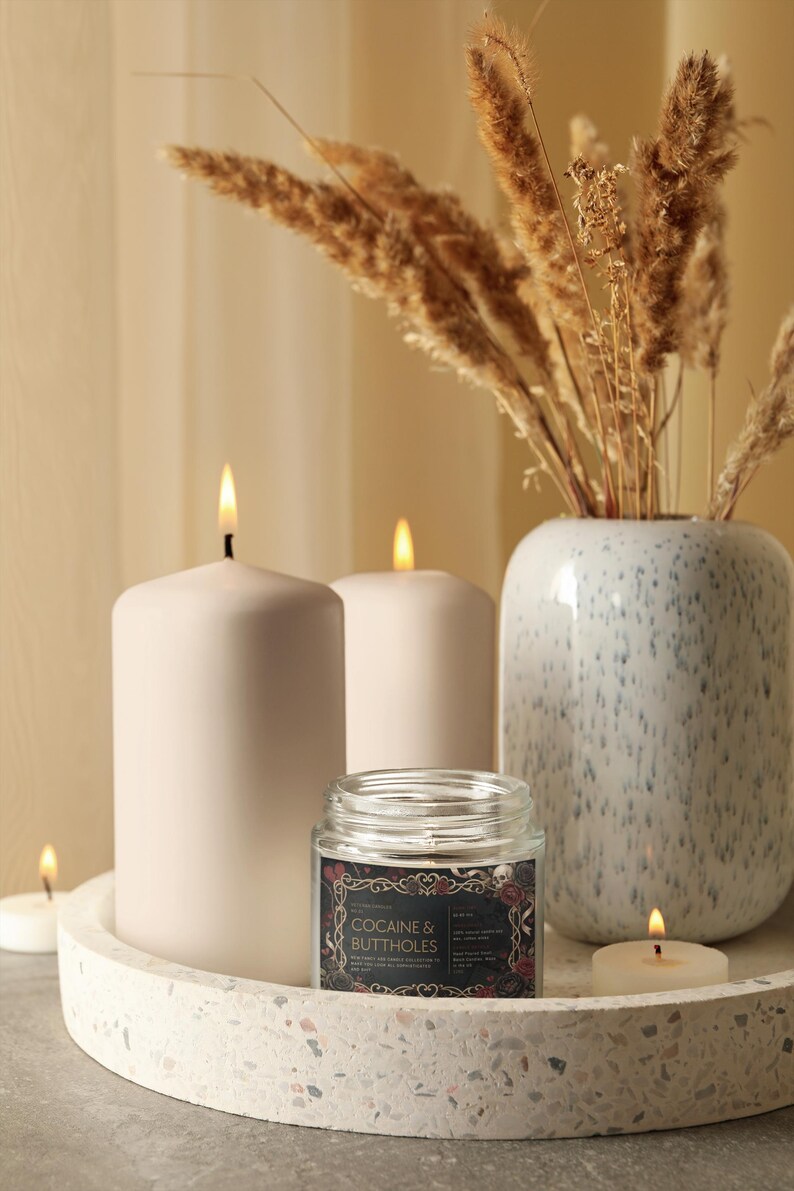 May include: A decorative arrangement featuring lit candles, a vase with dried plants, and a jar candle. The jar candle has a black label with the text "COCAINE & BUTTHOLES" and floral and skull graphics. The candles are white and sit on a round tray.