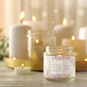May include: A lit glass jar candle with a floral label that reads "Cocaine & Buttholes" is the focal point. Several other lit candles of varying sizes and shapes are in the background, creating a warm, inviting ambiance. The candles are on a light-colored wooden surface.