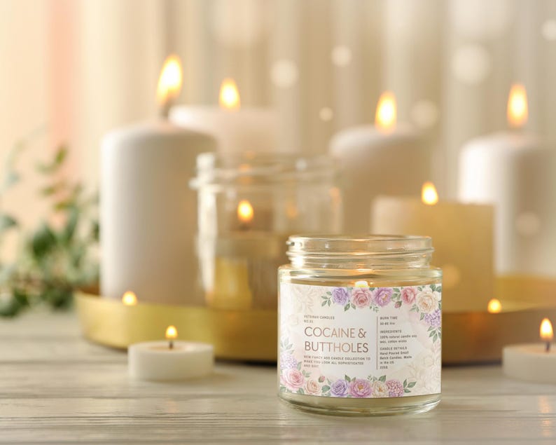 May include: A lit glass jar candle with a floral label that reads "Cocaine & Buttholes" is the focal point. Several other lit candles of varying sizes and shapes are in the background, creating a warm, inviting ambiance. The candles are on a light-colored wooden surface.