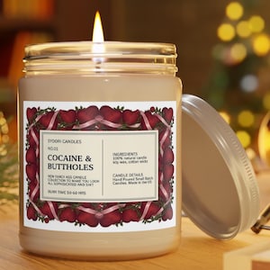 Puede incluir: Una vela encendida en un tarro de cristal con tapa plateada. La etiqueta dice "COCAINE & BUTTHOLES" con texto adicional. La etiqueta est&aacute; enmarcada con corazones rojos y cintas rosas. La vela est&aacute; sobre una superficie de madera.