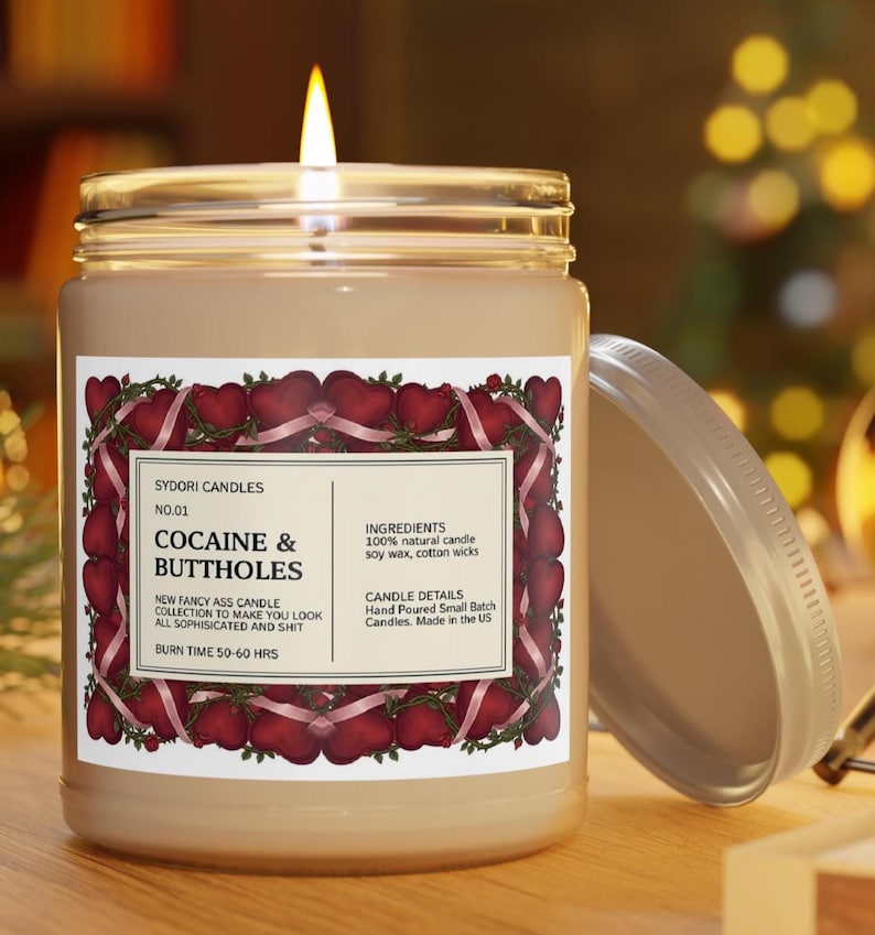 Puede incluir: Una vela encendida en un tarro de cristal con tapa plateada. La etiqueta dice "COCAINE & BUTTHOLES" con texto adicional. La etiqueta est&aacute; enmarcada con corazones rojos y cintas rosas. La vela est&aacute; sobre una superficie de madera.