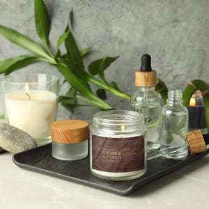 May include: A lit candle in a glass jar with a brown label that reads "COCAINE & BUTTHOLES" sits on a black tray. Other candles, glass bottles, and bamboo stalks are also on the tray, creating a spa-like aesthetic.