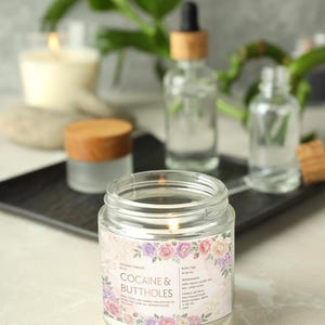May include: A lit glass jar candle with a floral label that reads "Cocaine & Buttholes." The label features a border of pink, purple, and white roses. Other glass bottles and a candle are in the background.