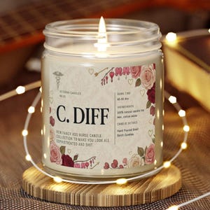 May include: A lit glass jar candle with a label featuring floral designs and the text "C. DIFF". The candle is on a wooden coaster, surrounded by fairy lights. The label includes the text "Veteran Candles" and "New Fancy Ass Nurse Candle Collection".