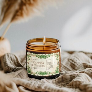 Puede incluir: Una vela encendida en un tarro de vidrio marr&oacute;n con una etiqueta que dice "Cocaine & Buttholes". La etiqueta tiene un borde floral verde y texto. La vela est&aacute; sobre una tela beige.