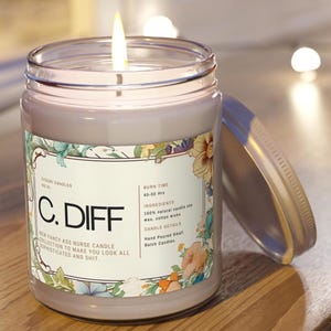 May include: A lit, lavender-coloured candle in a glass jar with a lid. The label reads "C. DIFF" and includes floral designs and text about the candle's ingredients and burn time. The candle is on a wooden surface.