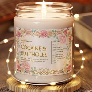 Puede incluir: Una vela encendida en un frasco de vidrio con una etiqueta que dice "COCAINE & BUTTHOLES". La etiqueta est&aacute; decorada con dise&ntilde;os florales y texto. La vela est&aacute; sobre un posavasos de madera, rodeada de luces de cadena.