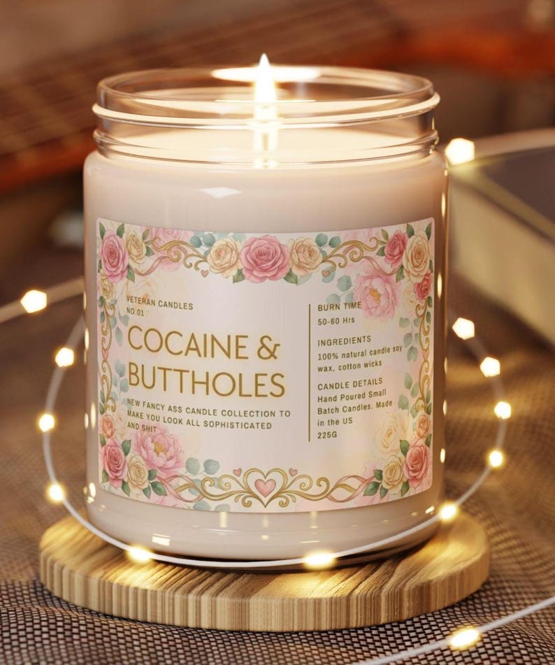 Puede incluir: Una vela encendida en un frasco de vidrio con una etiqueta que dice "COCAINE & BUTTHOLES". La etiqueta est&aacute; decorada con dise&ntilde;os florales y texto. La vela est&aacute; sobre un posavasos de madera, rodeada de luces de cadena.