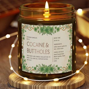 Może przedstawiać: Zapalone świeca w szklanym słoiku ze złotą obw&oacute;dką, na drewnianej podstawce. Etykieta głosi "COCAINE & BUTTHOLES" z dodatkowym tekstem. Świeca otoczona jest łańcuchem świetlnym.