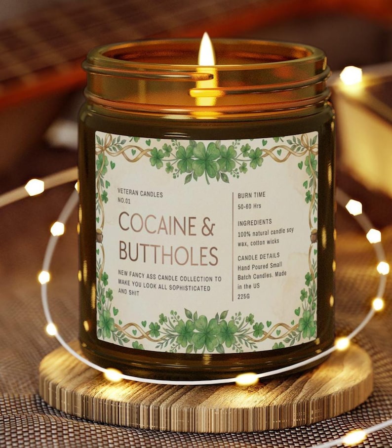 Może przedstawiać: Zapalone świeca w szklanym słoiku ze złotą obw&oacute;dką, na drewnianej podstawce. Etykieta głosi "COCAINE & BUTTHOLES" z dodatkowym tekstem. Świeca otoczona jest łańcuchem świetlnym.