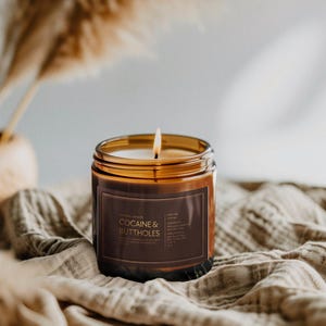 May include: A lit, amber-colored candle in a glass jar with a brown label that reads "COCAINE & BUTTHOLES." The candle sits on a beige cloth, with dried ornamental grass in the background. The candle's flame is visible.