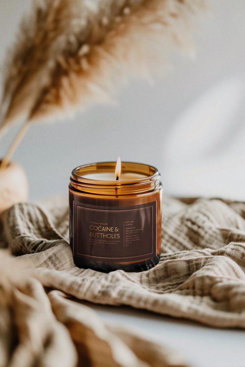 May include: A lit, amber-colored candle in a glass jar with a brown label that reads "COCAINE & BUTTHOLES." The candle sits on a beige cloth, with dried ornamental grass in the background. The candle's flame is visible.